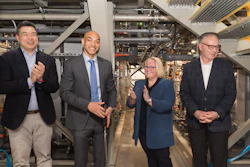 Four business professionals in formal attire standing together in an industrial facility with machinery and equipment visible in the background.