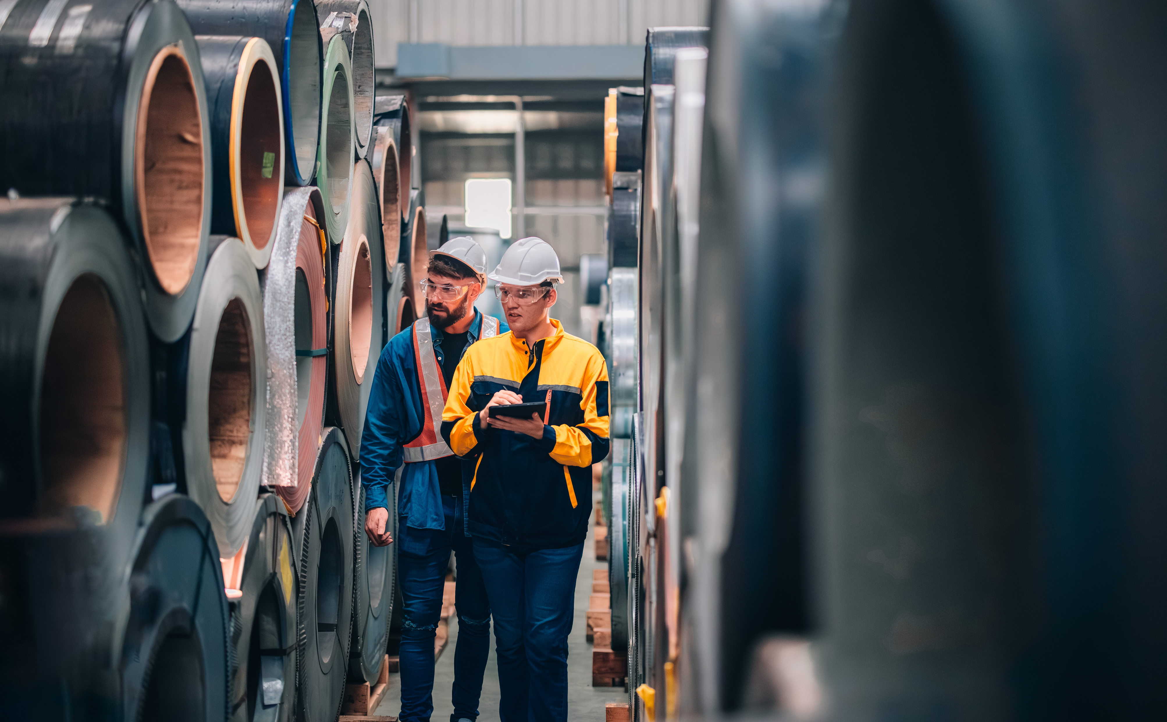 Two workers in safety gear inspecting rolled steel coils in an industrial warehouse facility