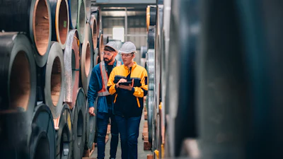 Two workers in safety gear inspecting rolled steel coils in an industrial warehouse facility
