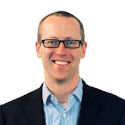 Professional headshot of smiling man wearing glasses, blue striped shirt, and black blazer