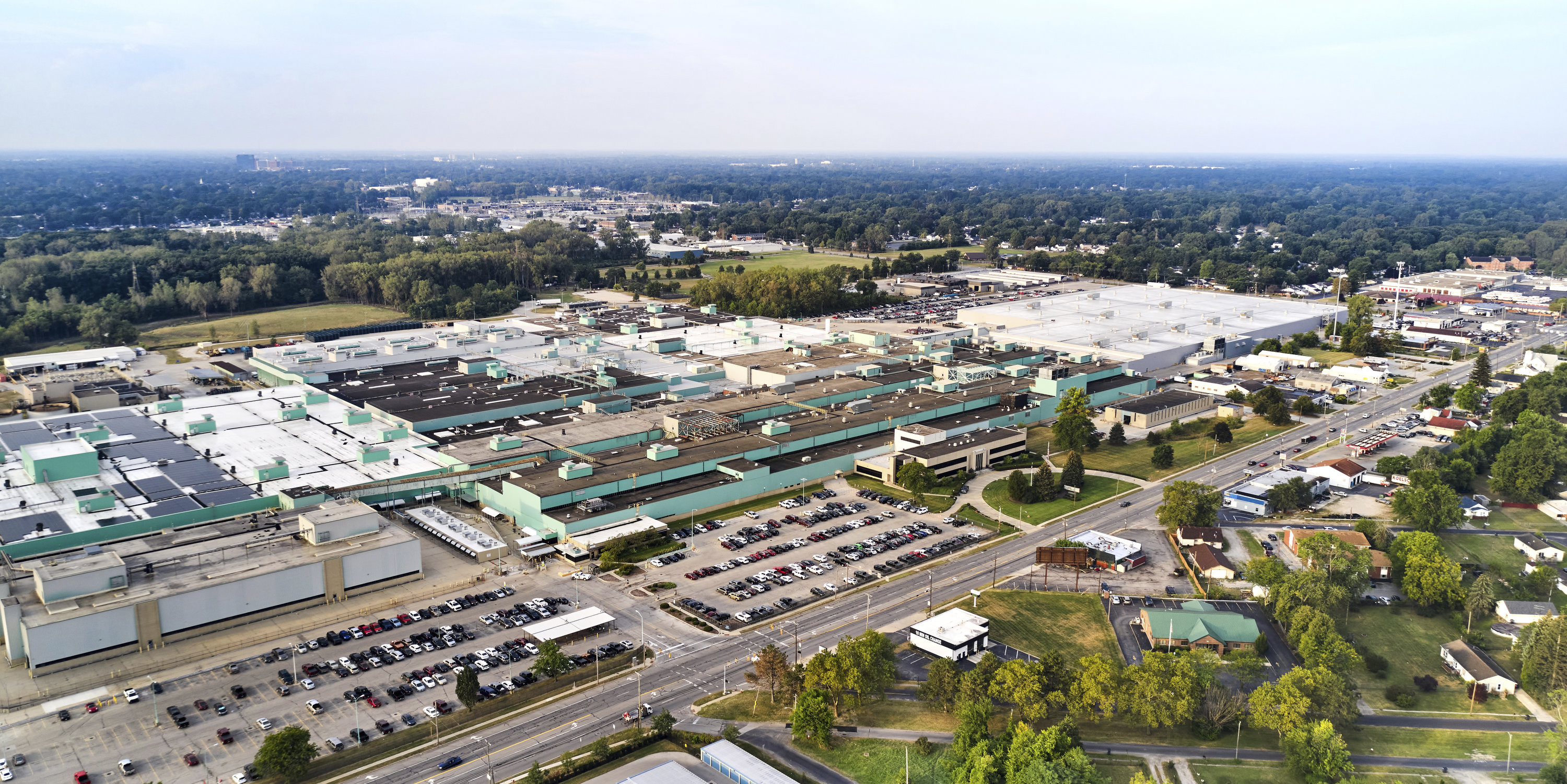 Aerial view of large industrial manufacturing facility with multiple green-roofed buildings, parking lots, and surrounding residential areas