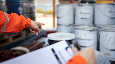 Worker in orange safety jacket pointing at stacked paint cans with clipboard in warehouse setting