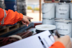 Worker in orange safety jacket pointing at stacked paint cans with clipboard in warehouse setting