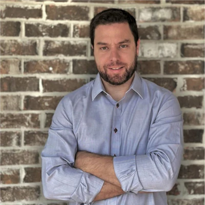 Professional portrait of a man with dark hair and beard wearing a light blue button-up shirt against a brick wall background
