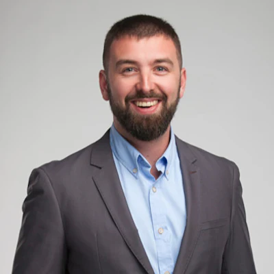 Professional headshot of a smiling man with beard wearing a gray blazer and light blue shirt