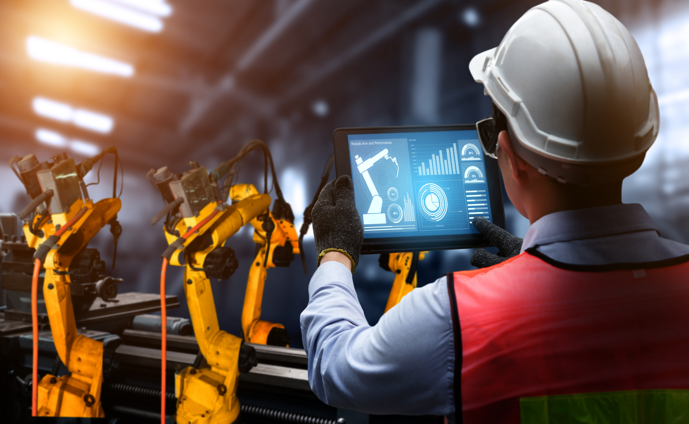Factory worker in hard hat monitoring industrial robotic arms with tablet displaying production analytics and performance metrics