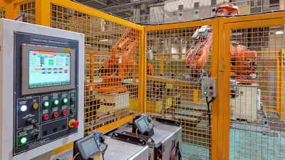 Industrial robotic assembly cell with orange safety enclosure and control panel in factory warehouse
