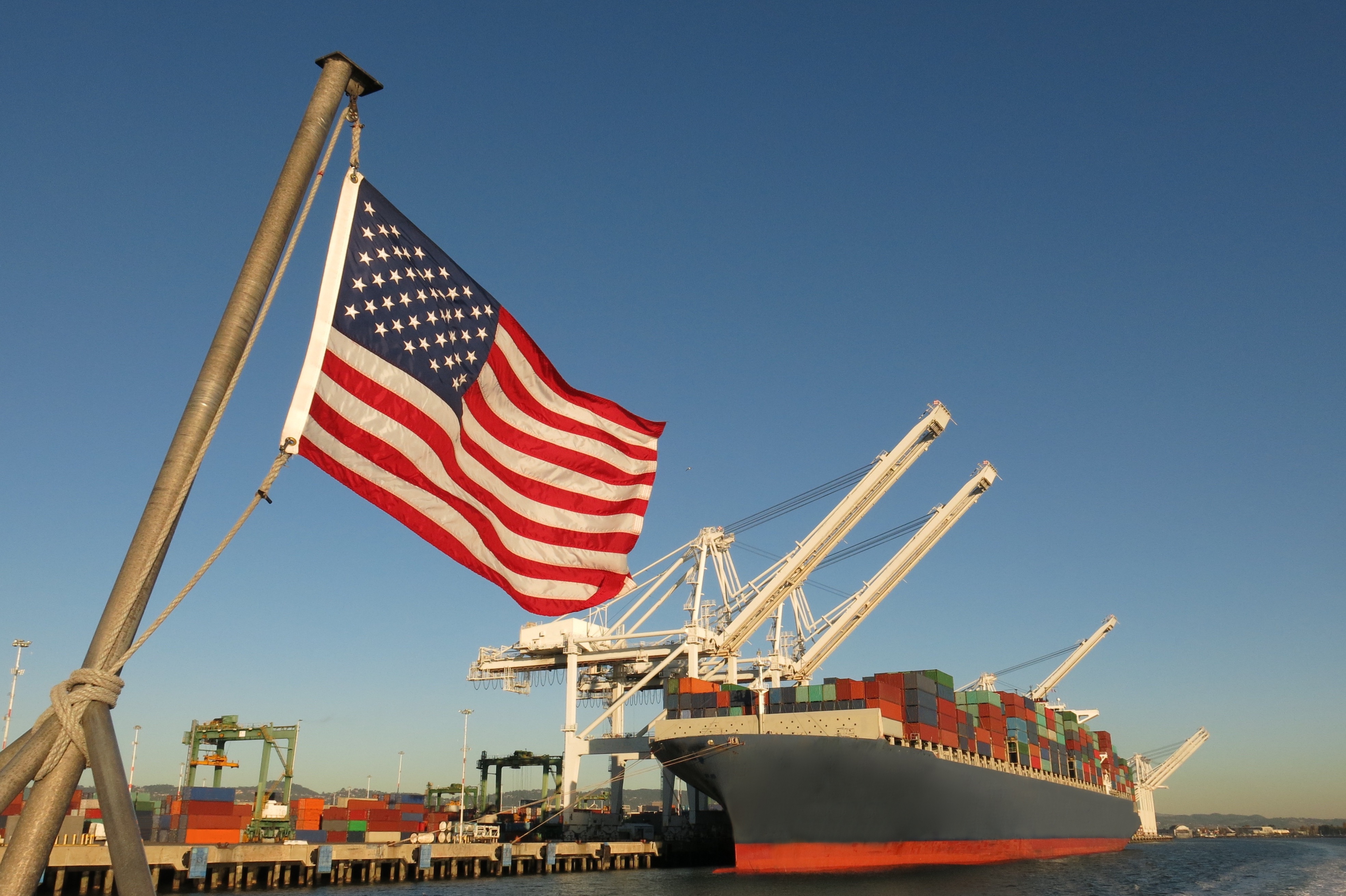 American flag flying at a cargo port with container ship and loading cranes against clear blue sky
