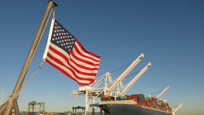 American flag flying at a cargo port with container ship and loading cranes against clear blue sky
