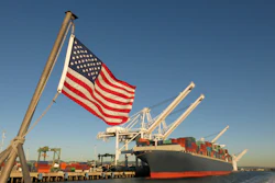 American flag flying at a cargo port with container ship and loading cranes against clear blue sky
