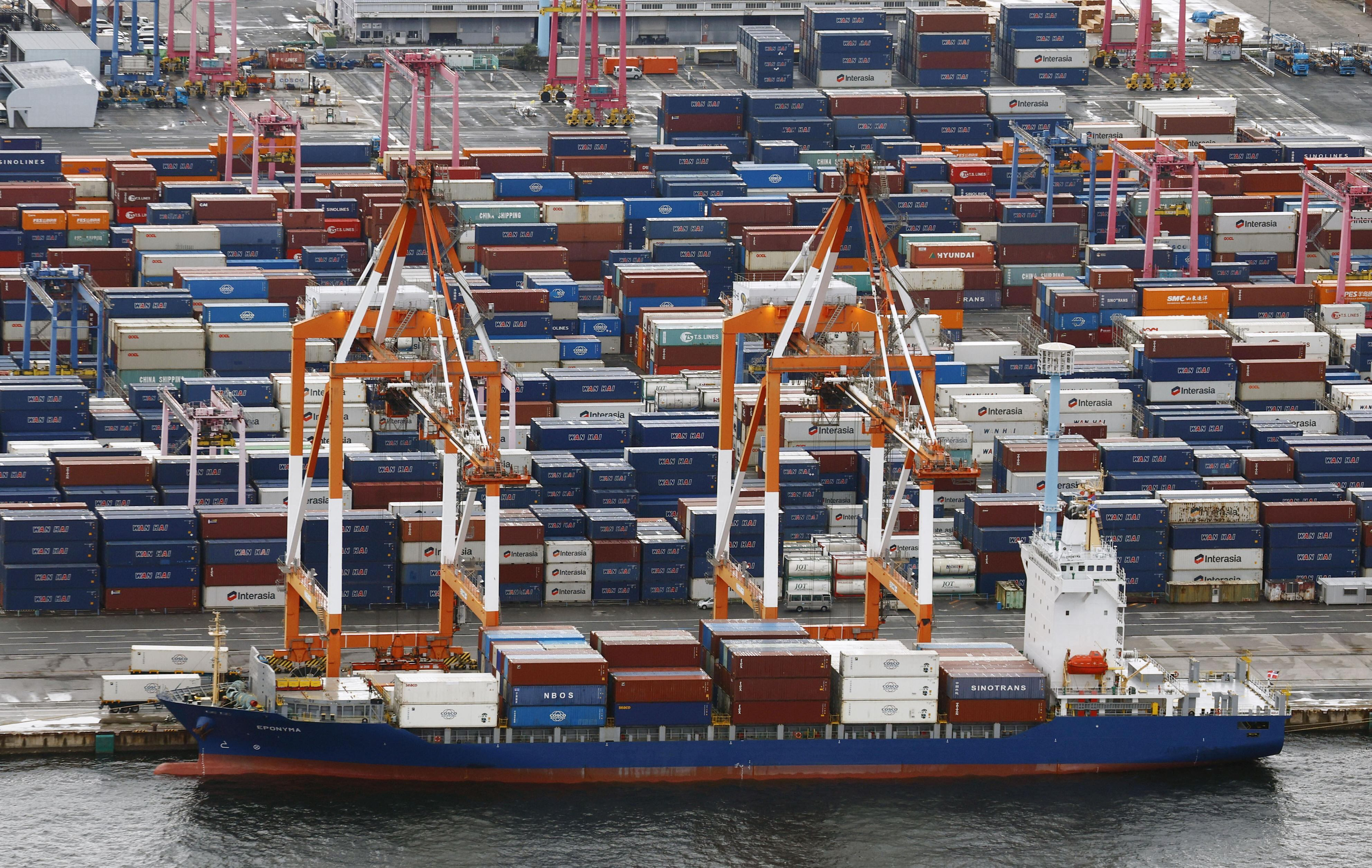 Cargo ship docked at container port with stacked shipping containers and orange loading cranes