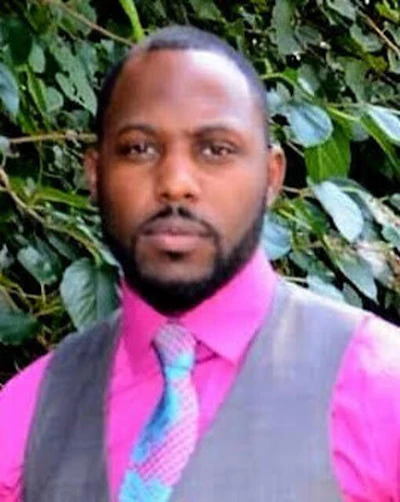 Professional portrait of a man wearing a bright pink shirt and gray vest against a green foliage background