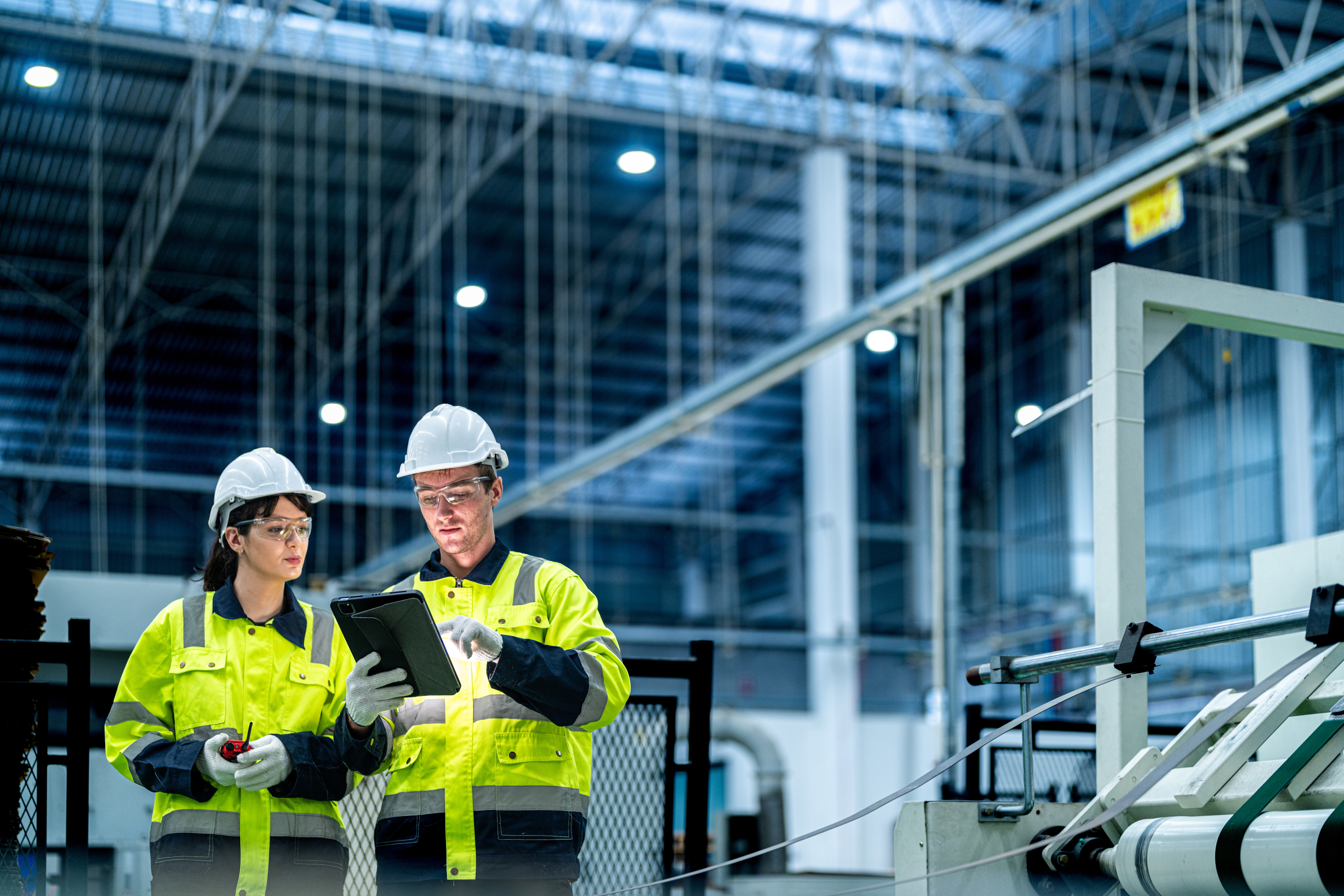 Two construction workers in hard hats and high-visibility vests review plans on a tablet at a modern building site.