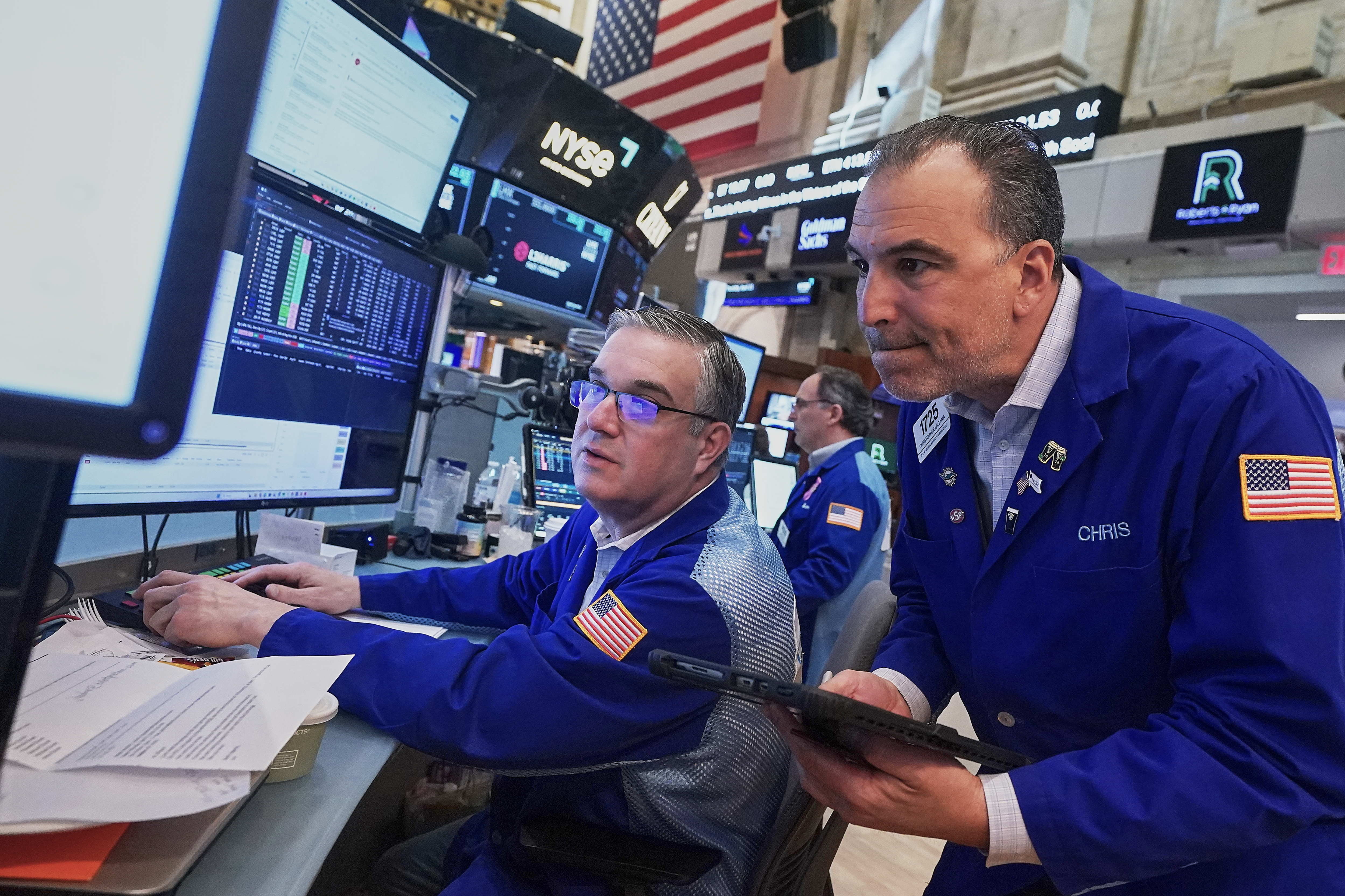 Traders monitor financial data at NYSE trading floor with multiple screens and American flags visible