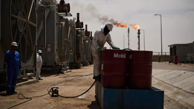Workers maintain industrial equipment at an oil refinery with gas flares burning in the background