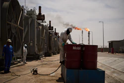 Workers maintain industrial equipment at an oil refinery with gas flares burning in the background