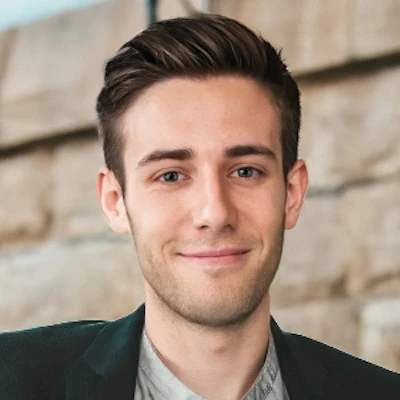 Professional headshot of a young man with dark hair wearing a dark blazer and gray shirt against a brick background