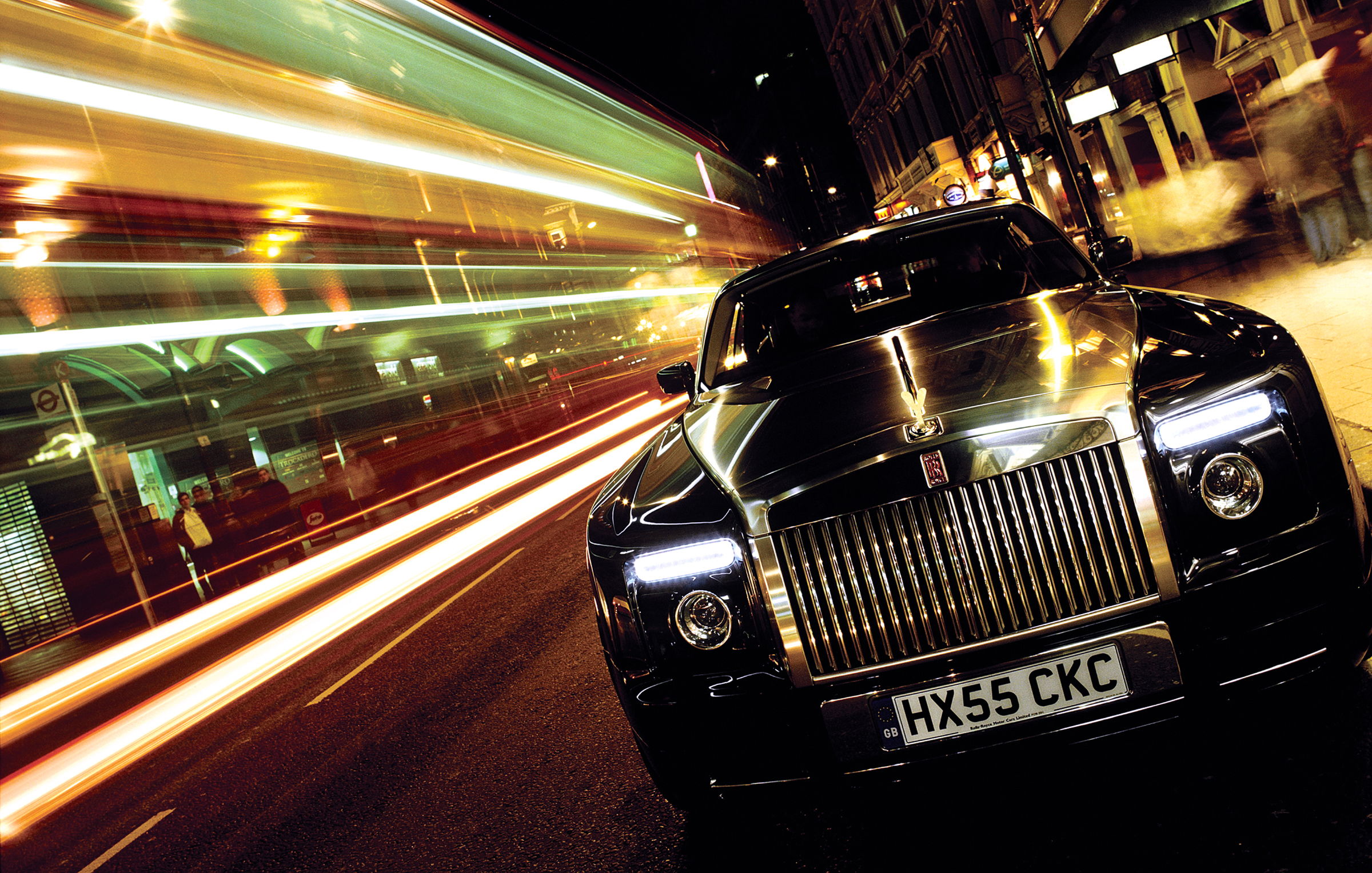 Black Rolls-Royce luxury car speeding through London street at night with motion-blurred bus and city lights