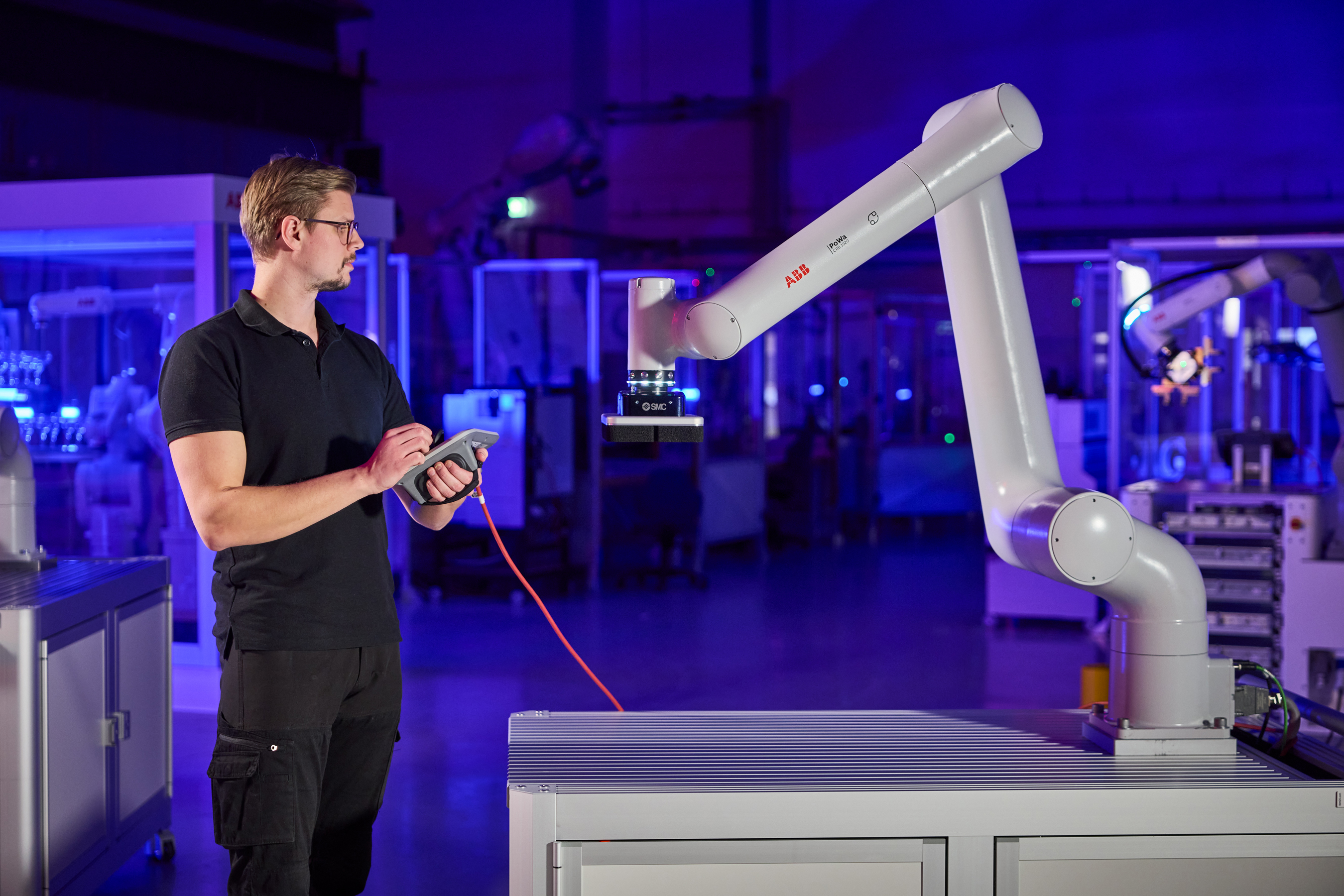 Technician operating white robotic arm with red controller in blue-lit industrial facility