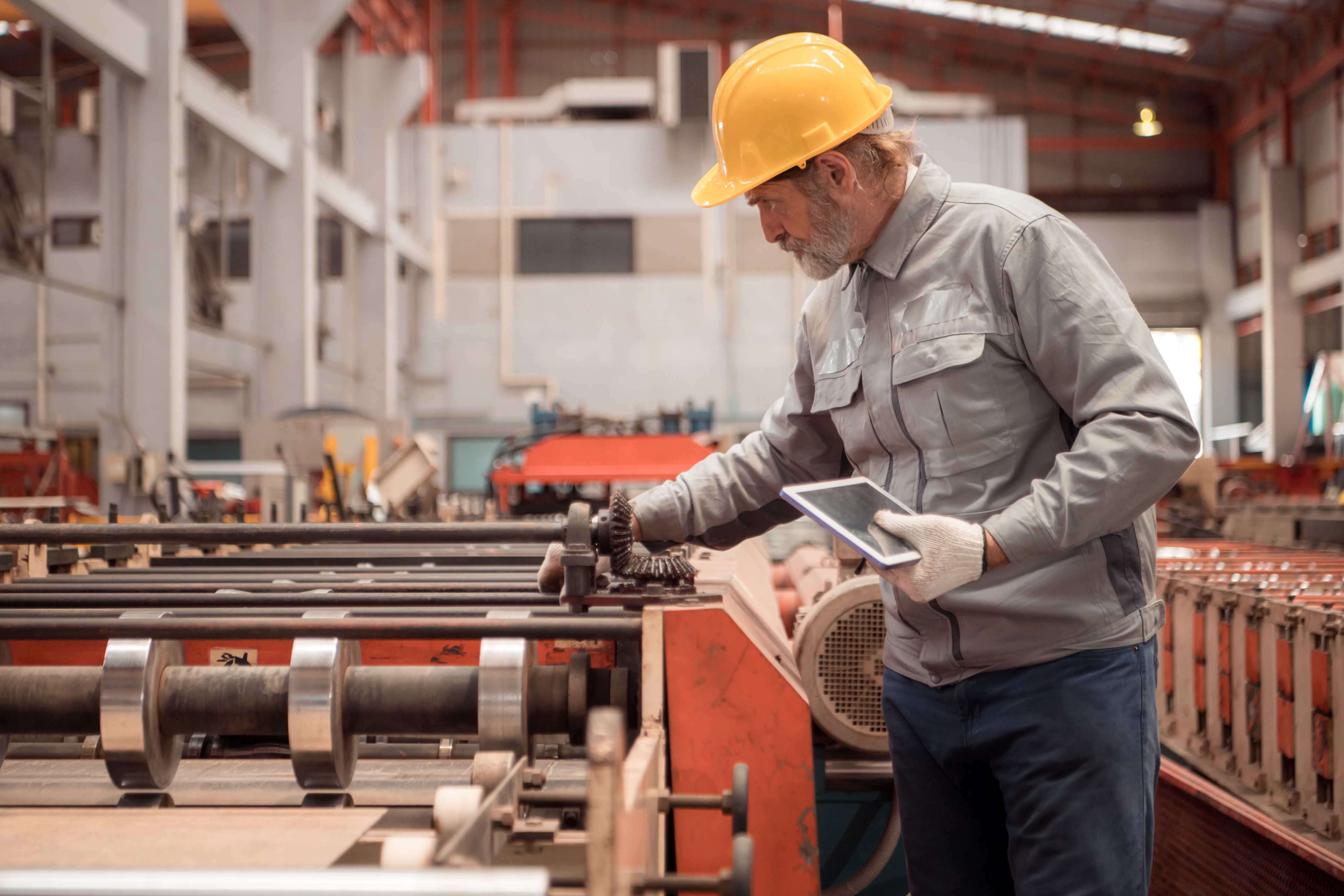 Experienced factory worker in hard hat reviewing tablet near industrial machinery in manufacturing plant