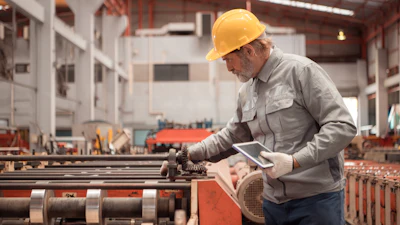 Experienced factory worker in hard hat reviewing tablet near industrial machinery in manufacturing plant