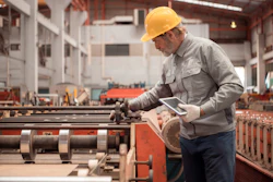 Experienced factory worker in hard hat reviewing tablet near industrial machinery in manufacturing plant