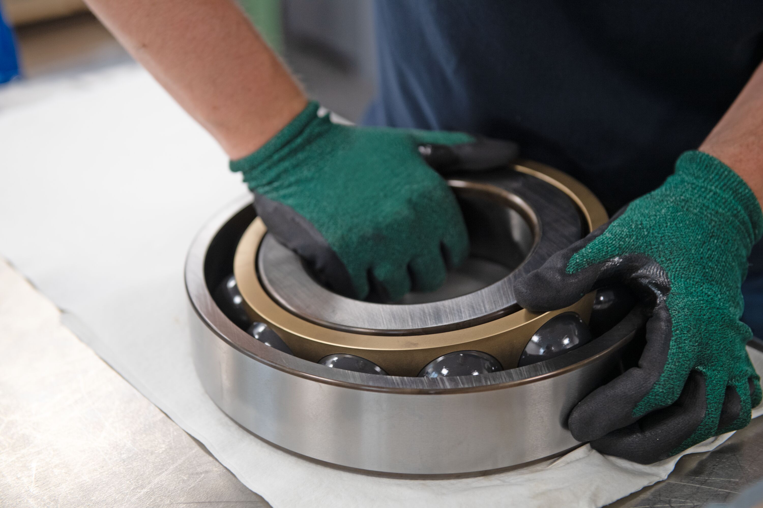 Worker in green gloves inspecting a large industrial ball bearing with metallic rings on white surface