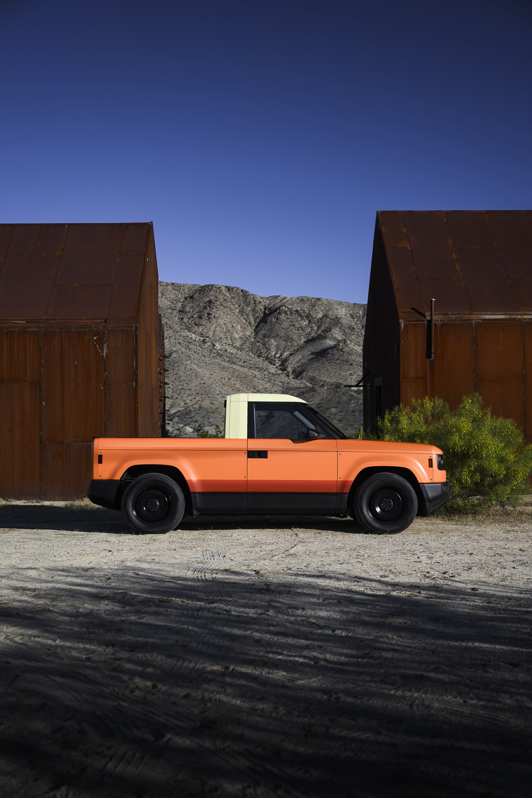 Orange pickup truck parked between rusted metal structures with desert mountains in background