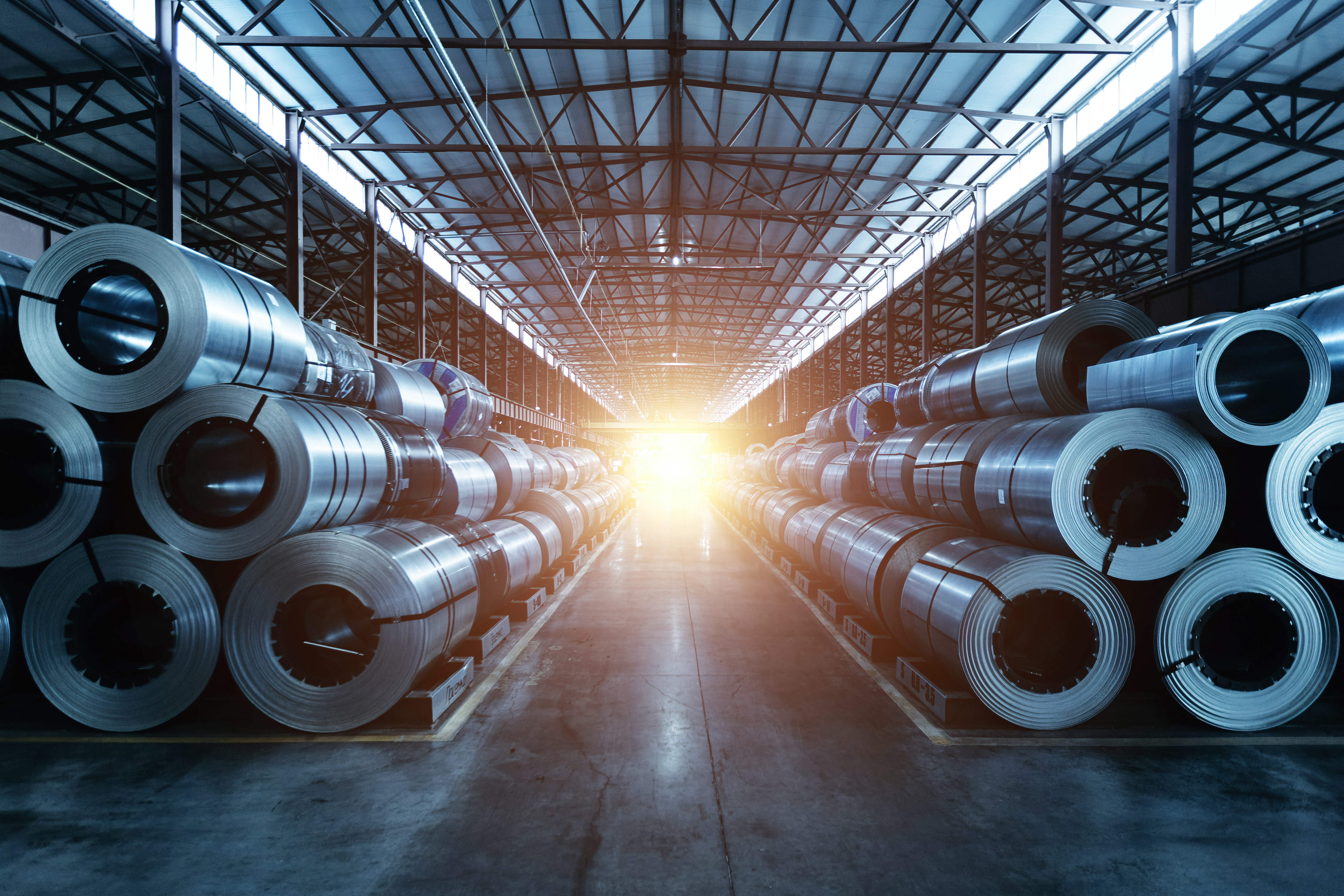 Industrial warehouse with stacked metal coils lining both sides of a central aisle under steel framework roof with natural light