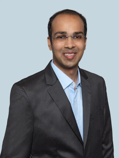 Professional headshot of a man wearing glasses and dark blazer over light blue shirt against neutral background