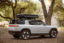 White Rivian R1S electric SUV with roof cargo box parked under large oak trees on gravel ground