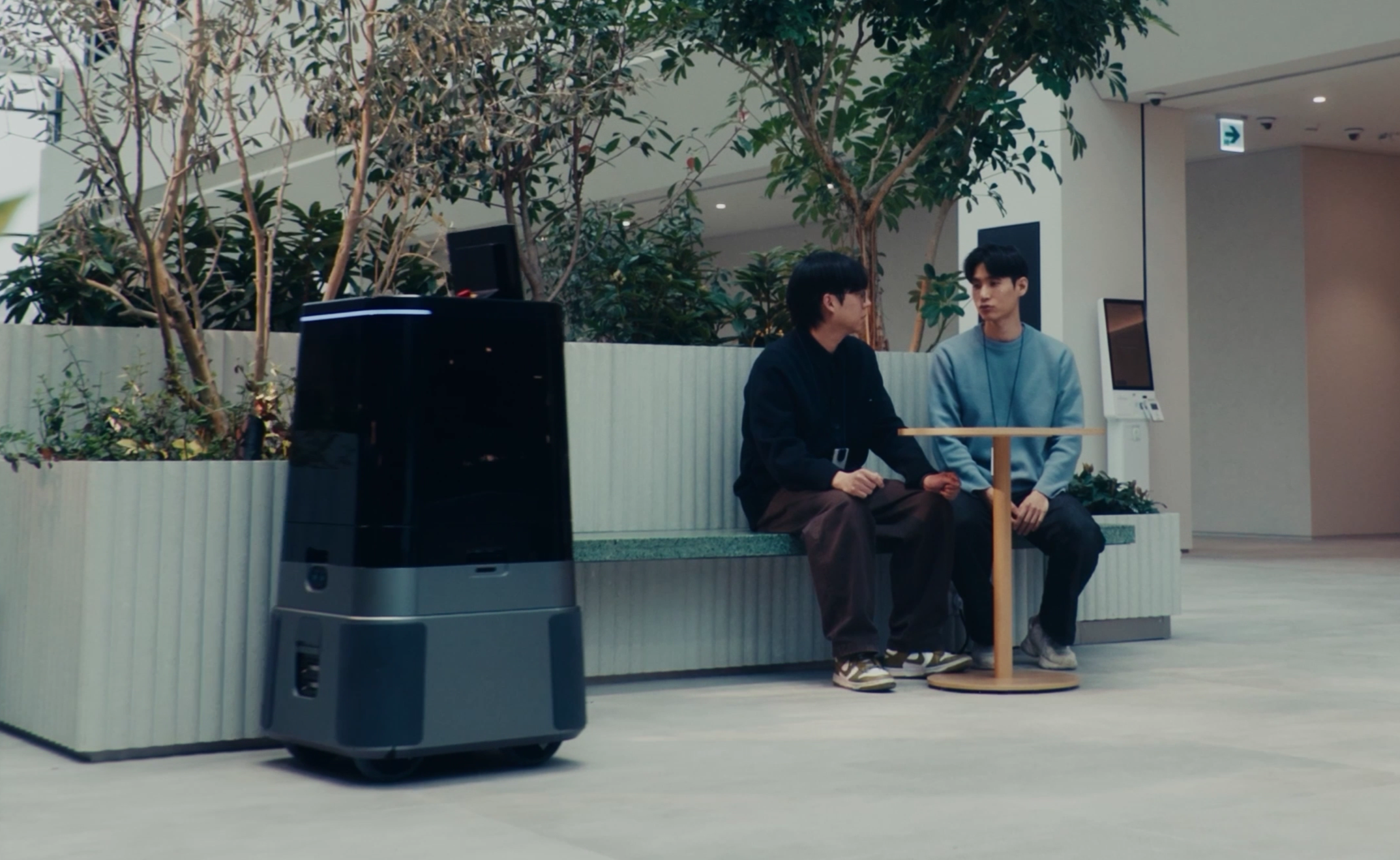 Two people sit on a bench in a modern office lobby with potted plants and a dark robotic device nearby.