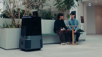 Two people sit on a bench in a modern office lobby with potted plants and a dark robotic device nearby.