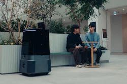 Two people sit on a bench in a modern office lobby with potted plants and a dark robotic device nearby.