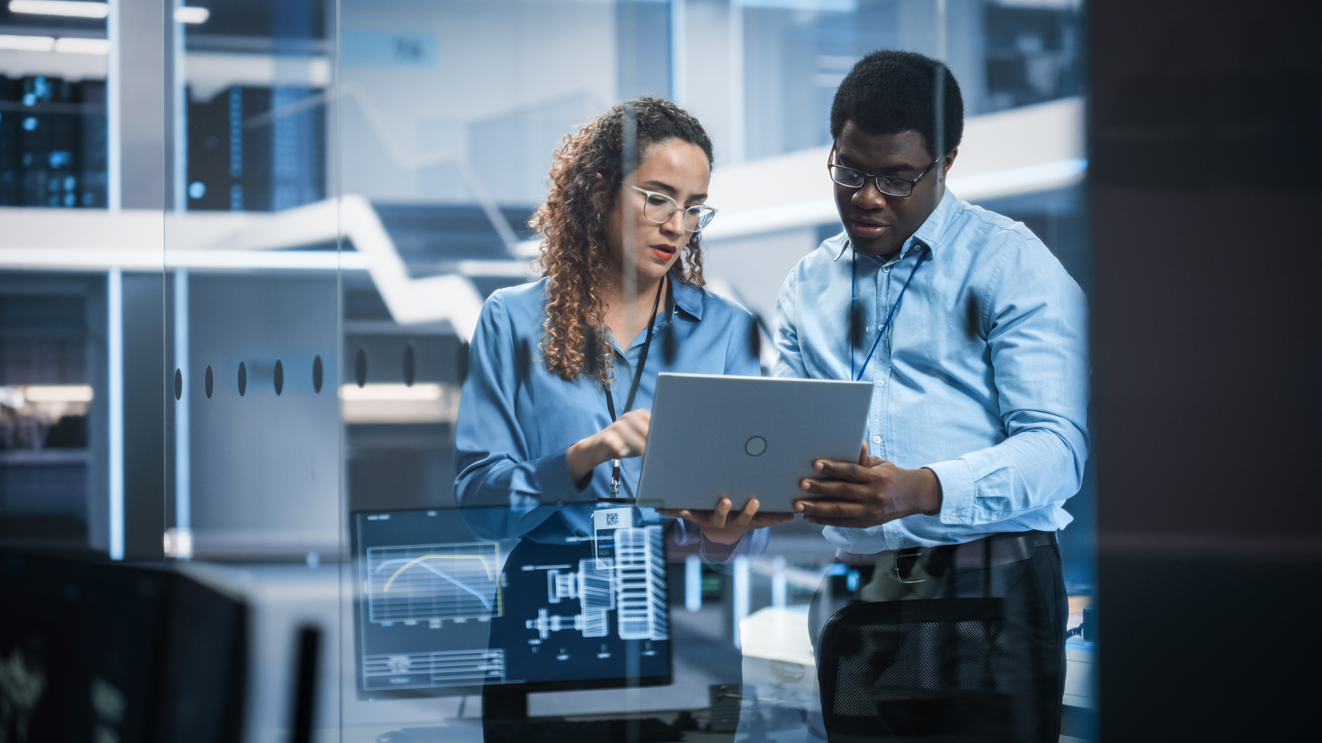 Two professionals in business attire collaborating on a laptop in a modern office environment with glass windows and digital displays.