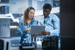 Two professionals in business attire collaborating on a laptop in a modern office environment with glass windows and digital displays.