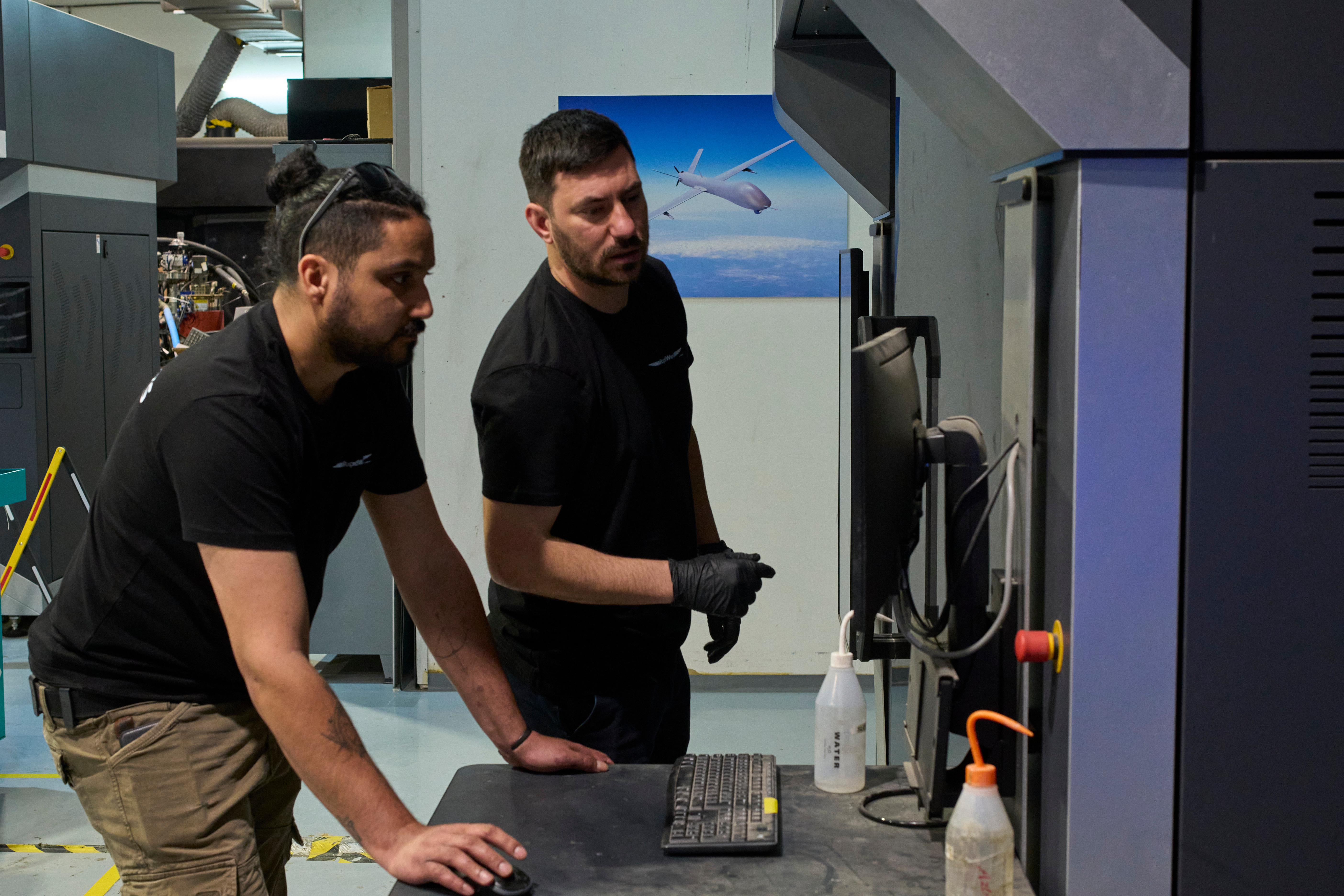 Two technicians in black shirts examining computer equipment and keyboard in a modern workspace with airplane poster on wall