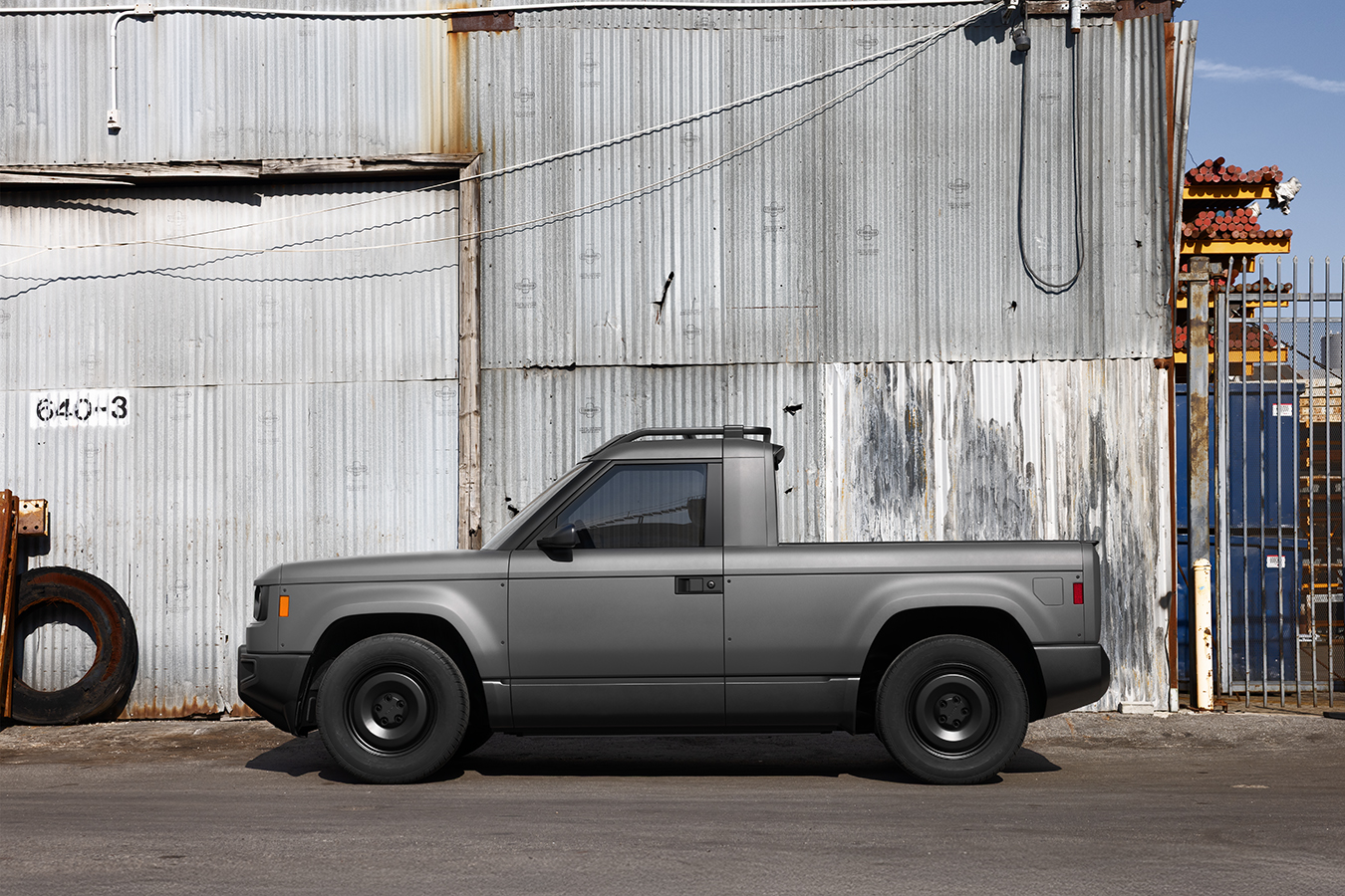 Gray compact pickup truck parked against industrial corrugated metal buildings with rusty equipment visible