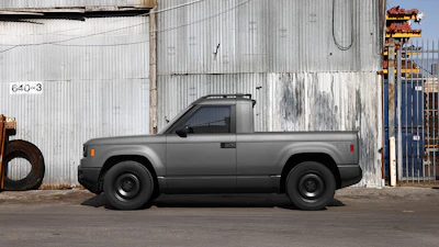 Gray compact pickup truck parked against industrial corrugated metal buildings with rusty equipment visible