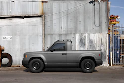 Gray compact pickup truck parked against industrial corrugated metal buildings with rusty equipment visible