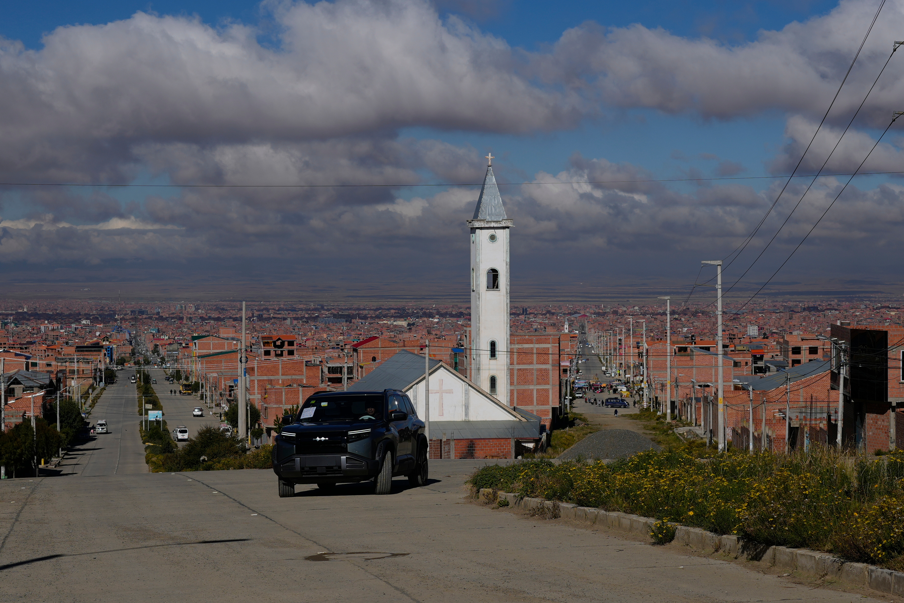 White church with tall steeple on hillside overlooking sprawling city with orange-brick buildings under cloudy sky