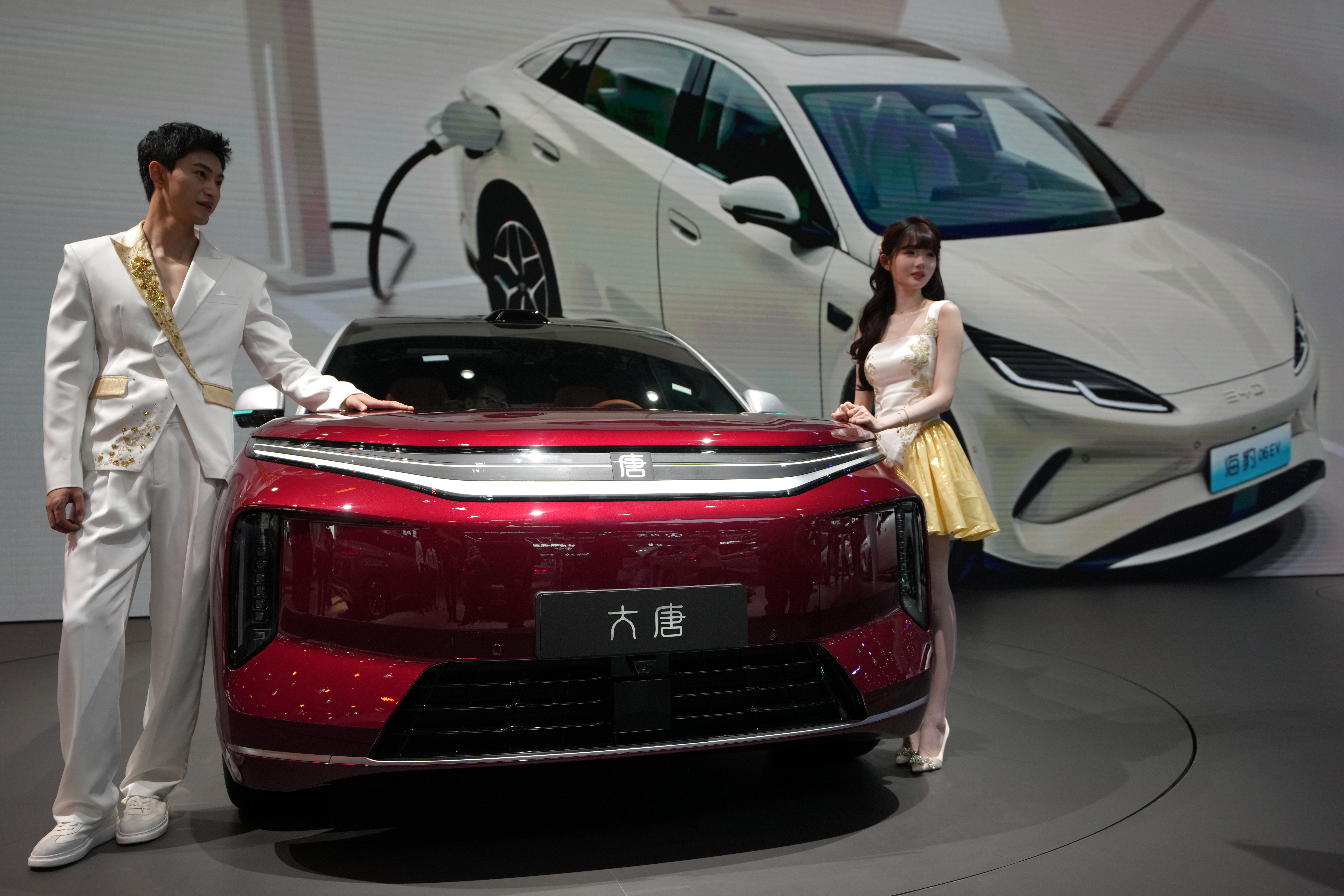 Two elegantly dressed hosts present a red electric vehicle alongside white EVs at a modern automotive showroom event.