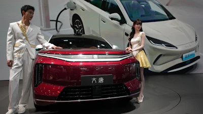 Two elegantly dressed hosts present a red electric vehicle alongside white EVs at a modern automotive showroom event.