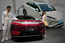 Two elegantly dressed hosts present a red electric vehicle alongside white EVs at a modern automotive showroom event.