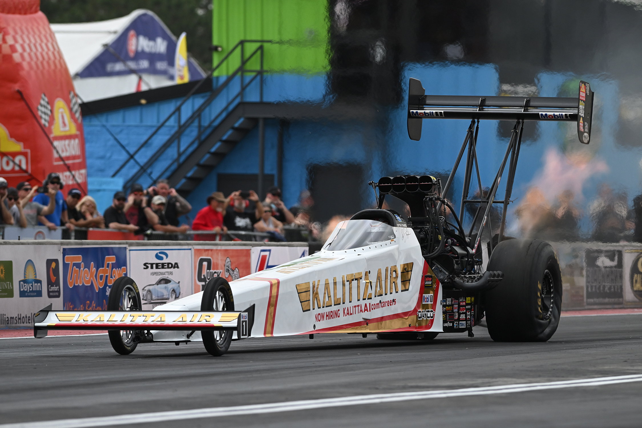 White and orange top fuel dragster racing with parachute deployed, smoke billowing, spectators watching from bleachers at motorsports event