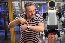 Man in striped shirt demonstrating white robotic device in industrial manufacturing facility
