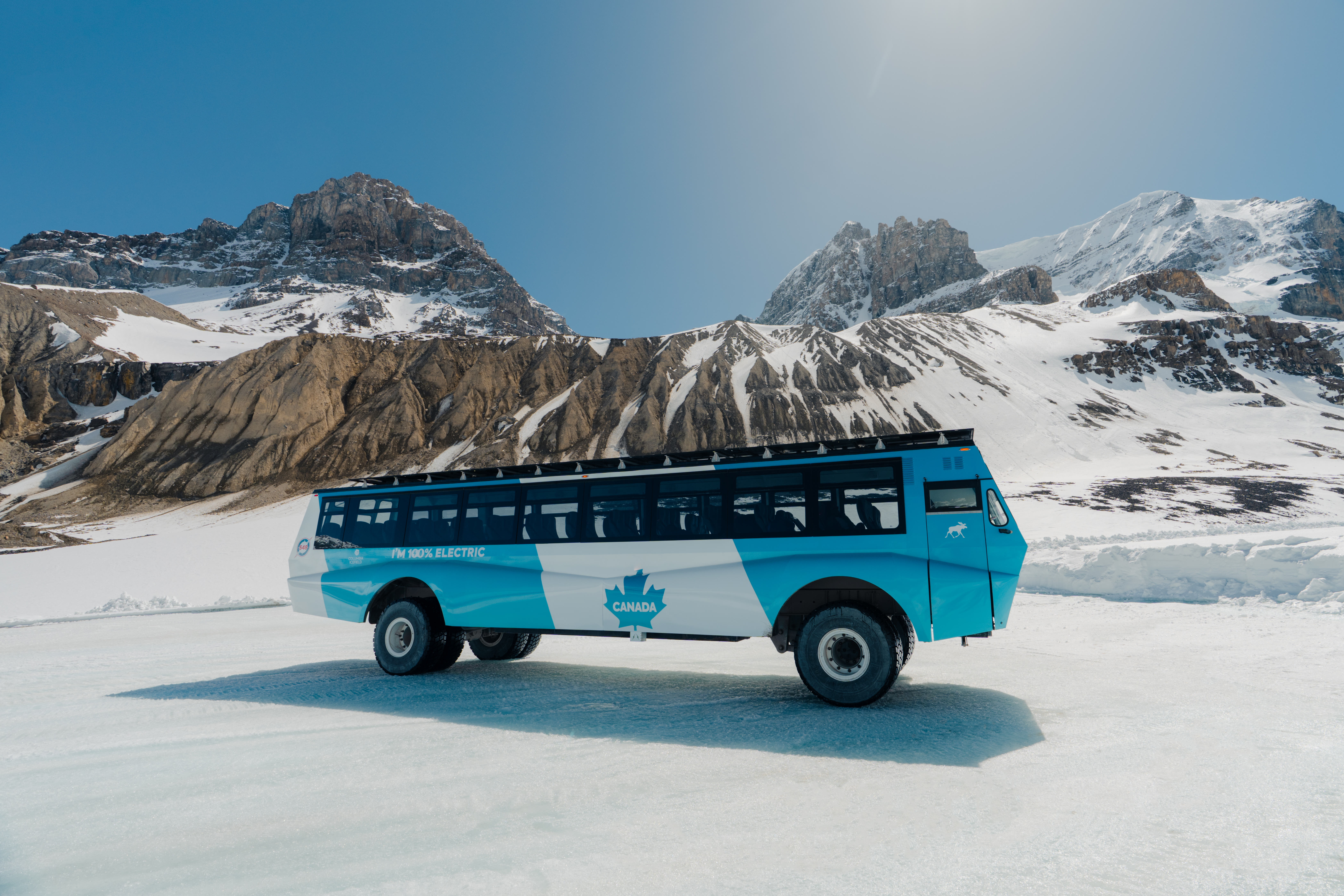 Turquoise tour bus with maple leaf emblem parked on frozen lake surrounded by snow-capped mountains under clear blue sky