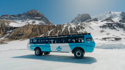 Turquoise tour bus with maple leaf emblem parked on frozen lake surrounded by snow-capped mountains under clear blue sky
