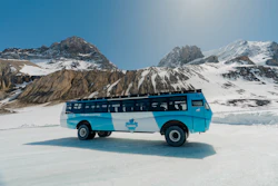 Turquoise tour bus with maple leaf emblem parked on frozen lake surrounded by snow-capped mountains under clear blue sky