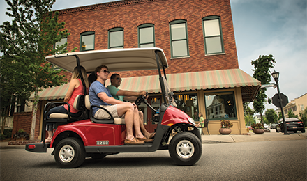 Family enjoying a red golf cart ride on a tree-lined street with brick buildings